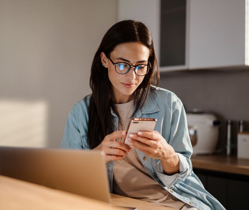 Soustředěná žena s brýlemi si prochází informace na smartphonu, s notebookem v pozadí, v domácí kanceláři.