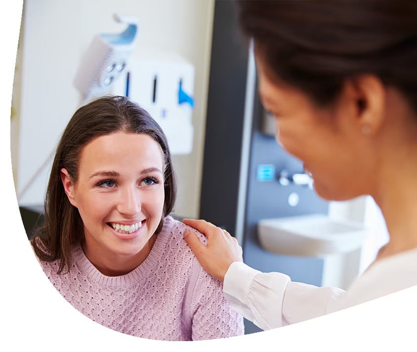 Usmívající se těhotná žena stojí u okna, drží se za břicho.