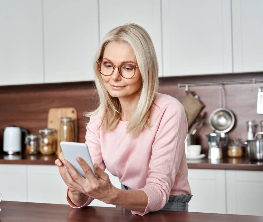 Starší žena v růžovém svetru čte zprávy na smartphonu v moderní kuchyni.